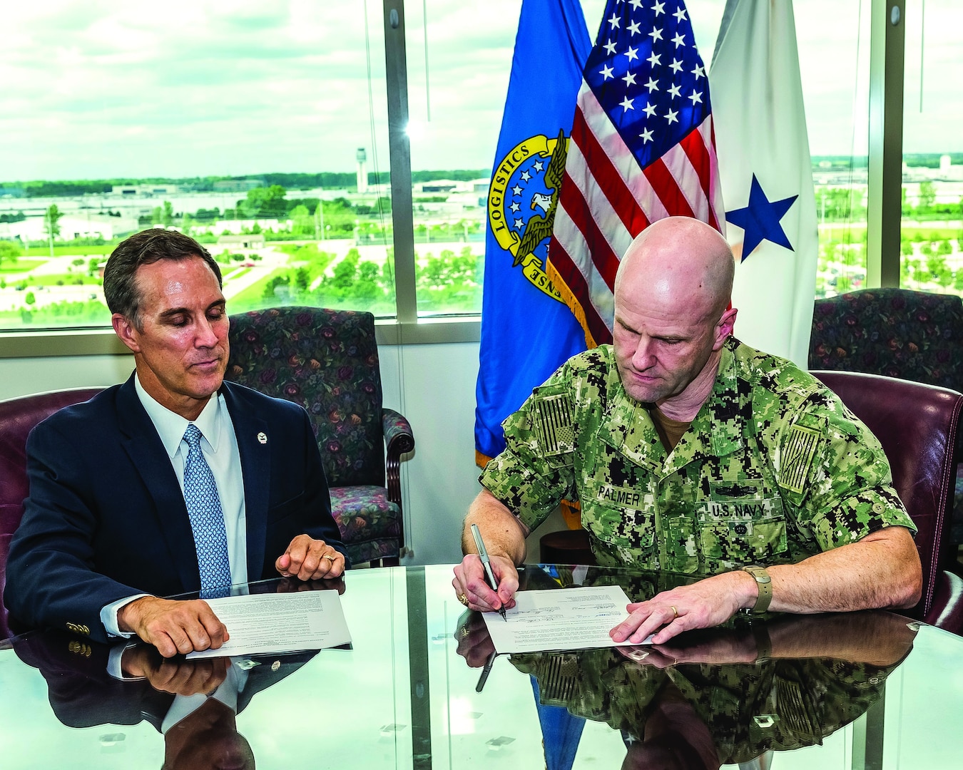 A civilian and military man sit at a table with papers in front of them.