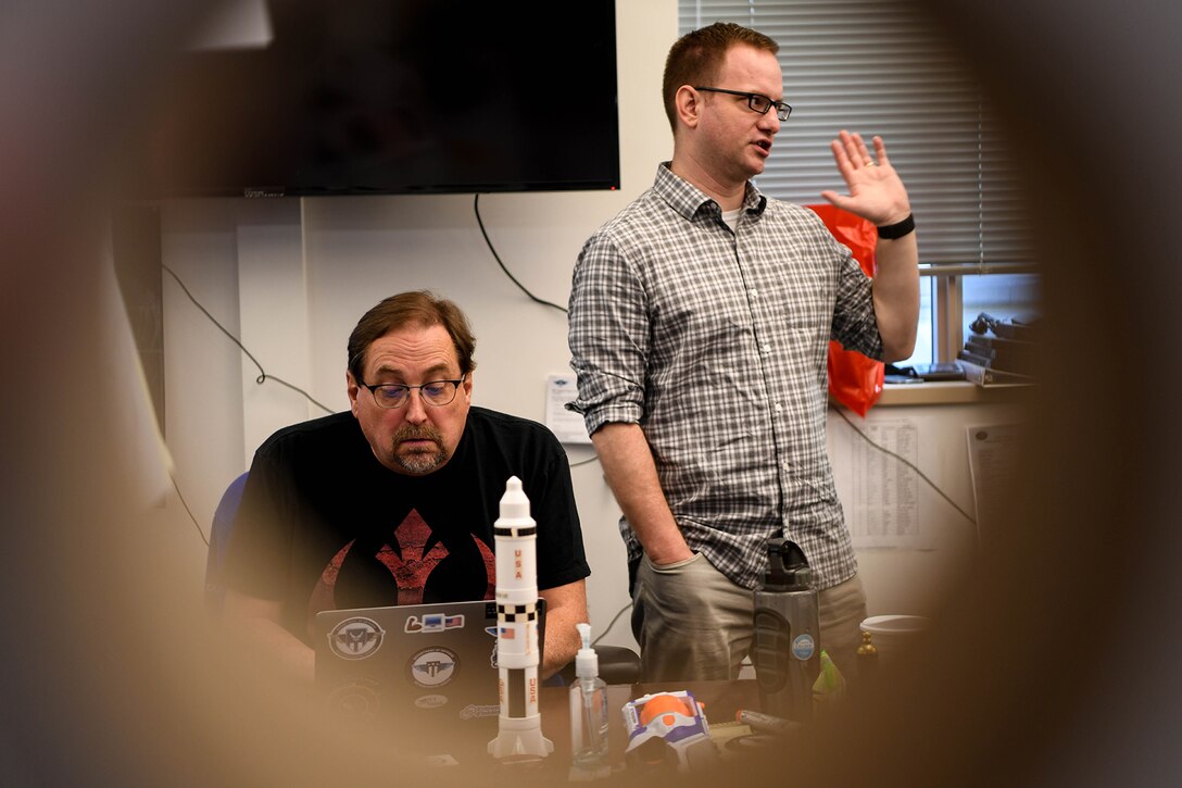 One man sitting down looking at his computer screen and another man standing up talking.