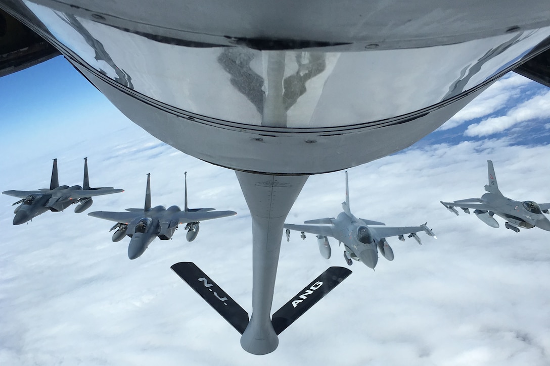 Four military jets fly in formation behind a bigger military aircraft.