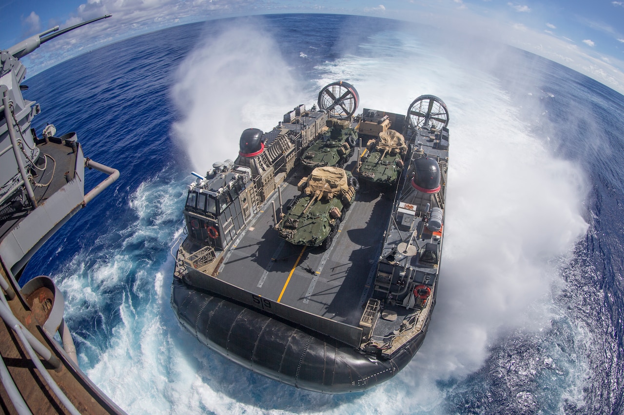 A boat carrying military vehicles splashes into the water.