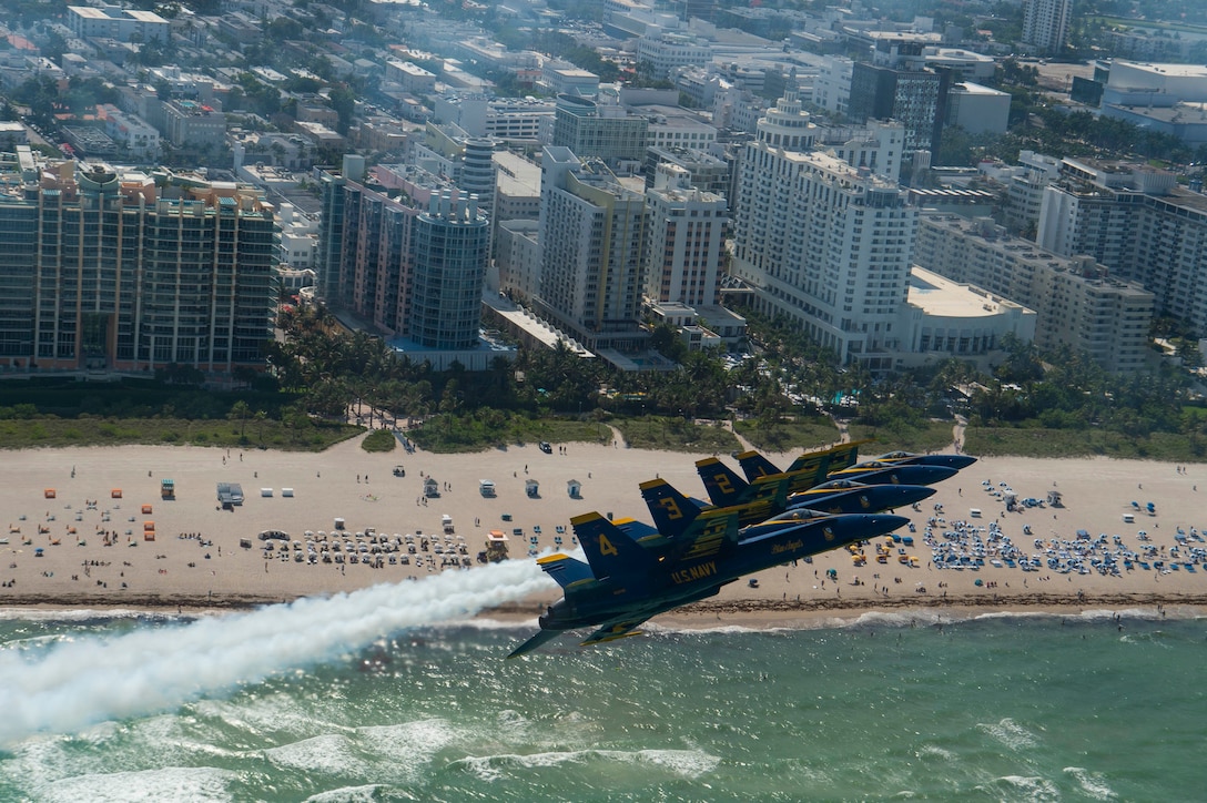 Four fighter jets fly in formation over the shoreline as spectators watch from the beach far below.