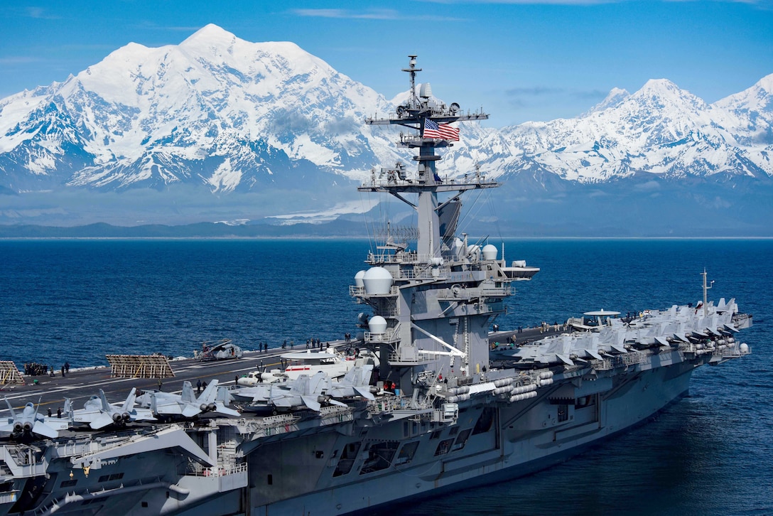 An aircraft carrier with several jets on its flight deck crosses the water as snowcapped mountains loom in the background.