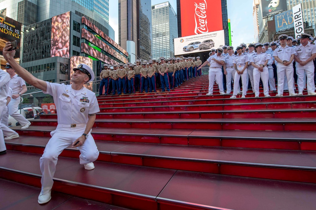 A sailor takes a selfie with a group of service members standing on red steps in front of billboards and buildings.