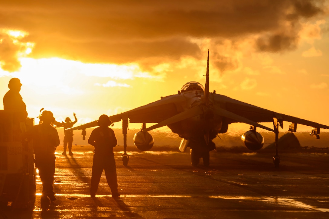 Sailors and Marines, shown in silhouette, stand and motion on a flight deck around an aircraft, against an orange sky.