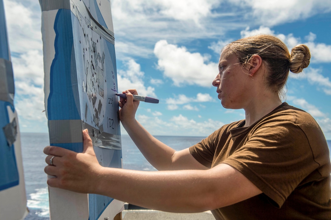 A sailor writes numbers on a target on a ship's flight deck, with blue sky in the background.