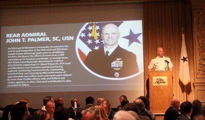 Navy member in dress whites stands on stage in front of a crowded room with a power point in the background