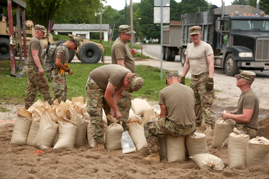 About seven soldiers pack sandbags near construction vehicles.