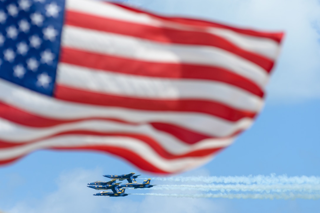Five fighter jets – some upside down – leave a trail across the sky as an American flag flies in the foreground.