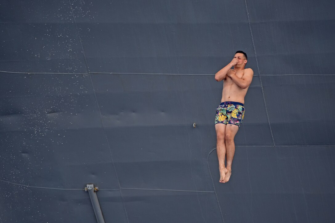 A sailor holds his breath and pinches his nose as he jumps feet-first into the water off the side of a ship.