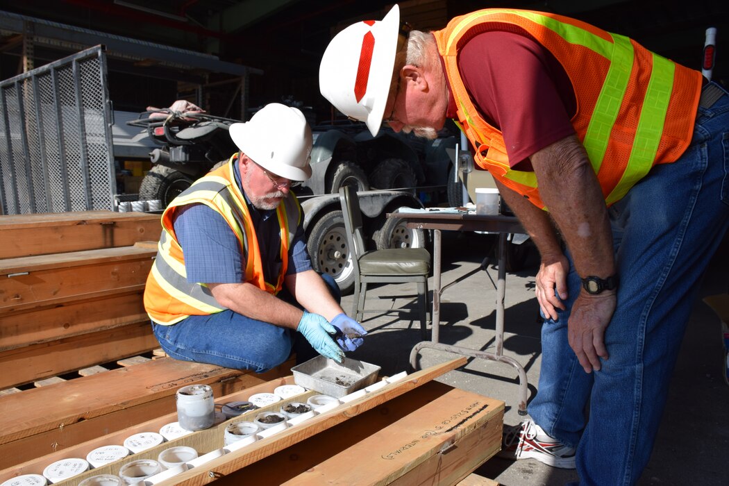 one employee sitting down and another one leaning to look at containers in a wooden crate