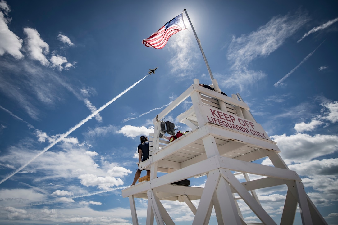 An Air Force fighter jet leaves a trail in the sky high above a lifeguard chair and an American flag.