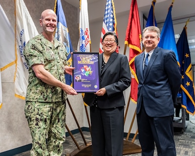 Event guest speaker being presented a memento of appreciation by two members of DLA Land and Maritime command staff.