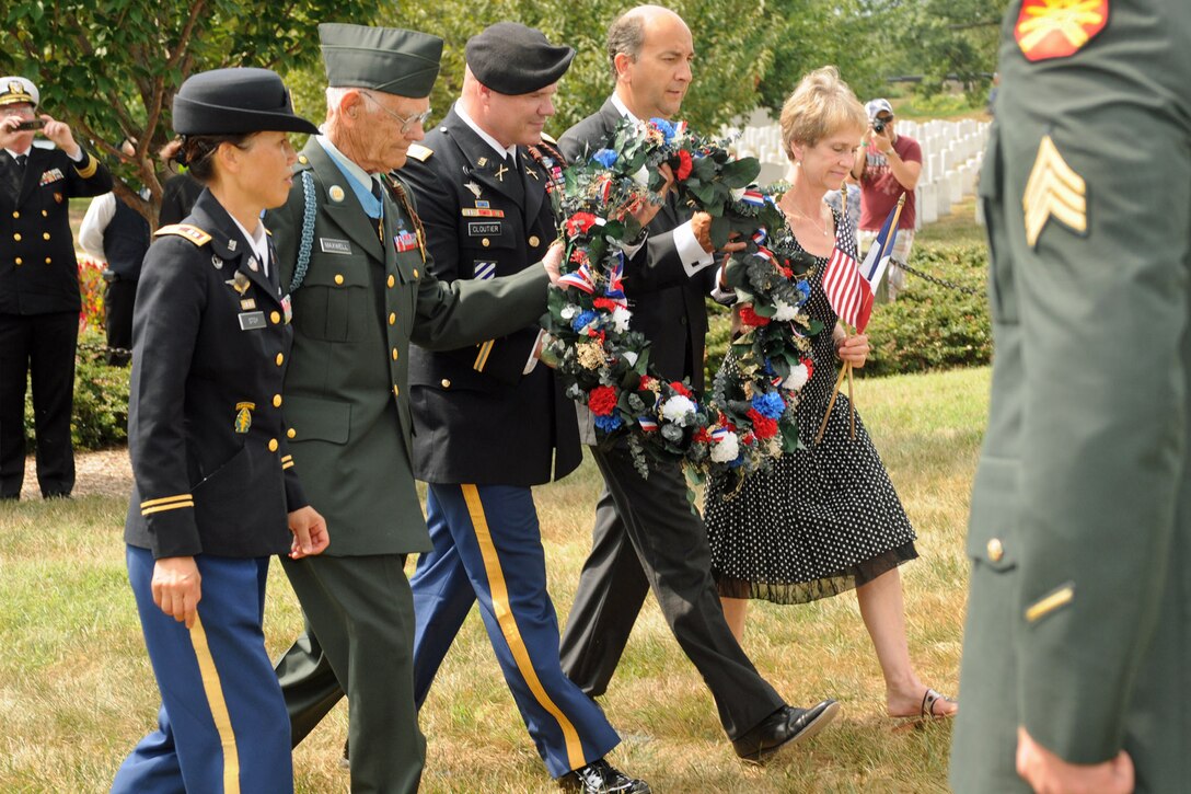 Four uniformed military members and a woman walk together in a cemetery while one of them holds a wreath.