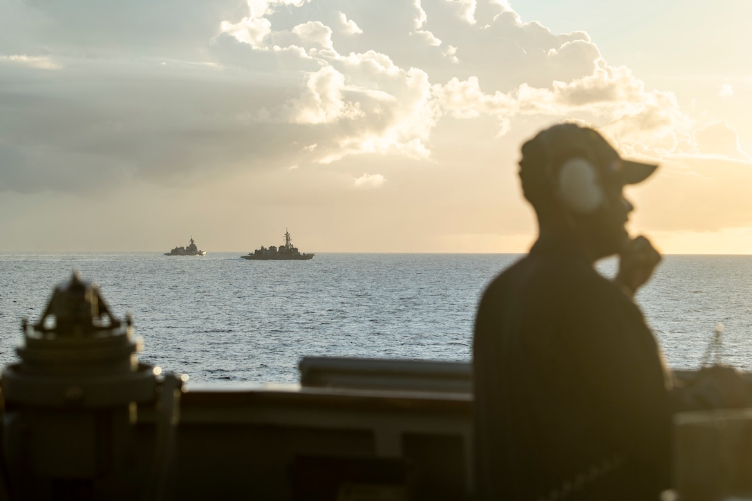 A sailor looks out to sea from a ship at twilight as two other military ships float nearly.