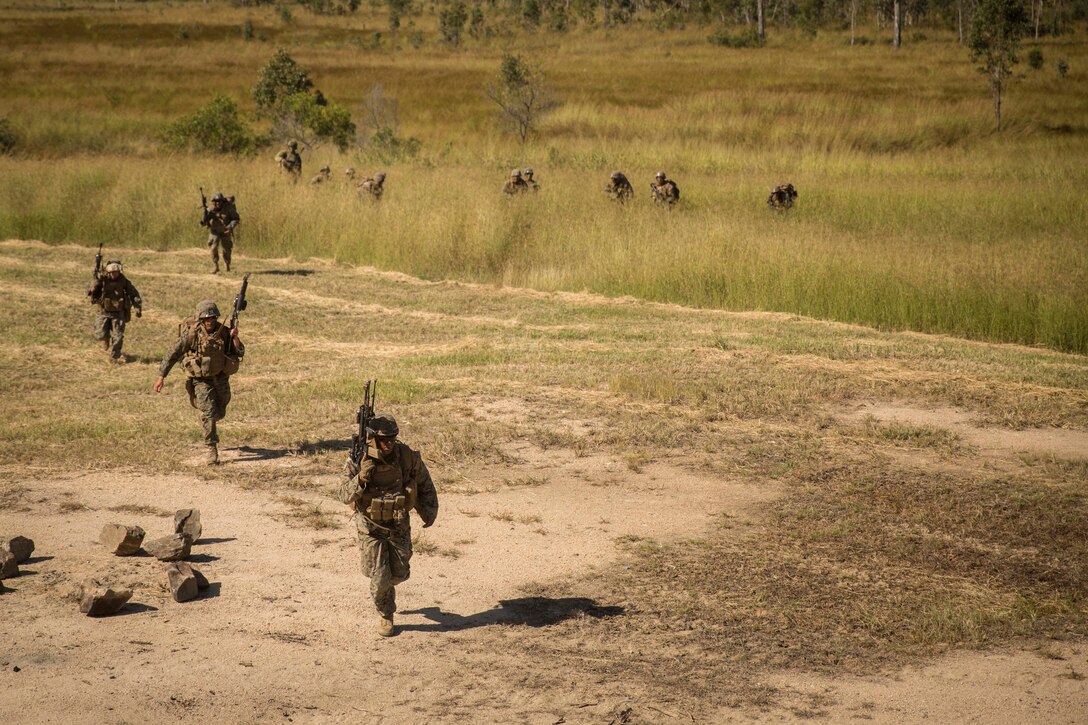 Thirteen Marines, wearing camouflage uniforms, vests and helmets and carrying weapons, walk from dense grassland past a clearing with a fire pit.