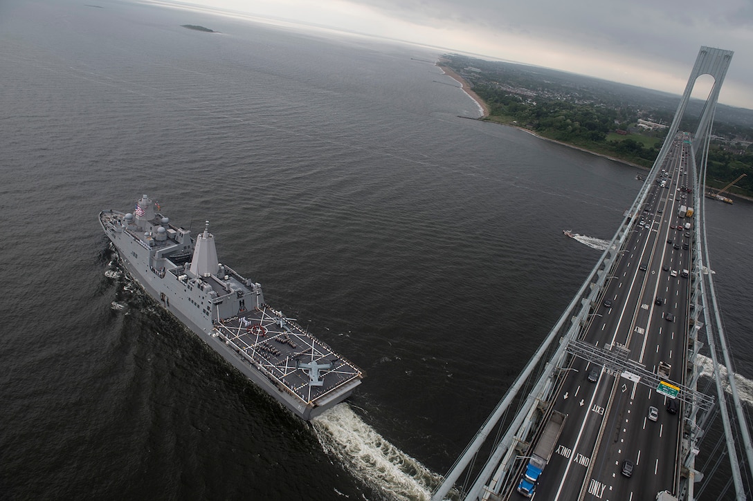 A military ship steams under a bridge as it heads out to sea.