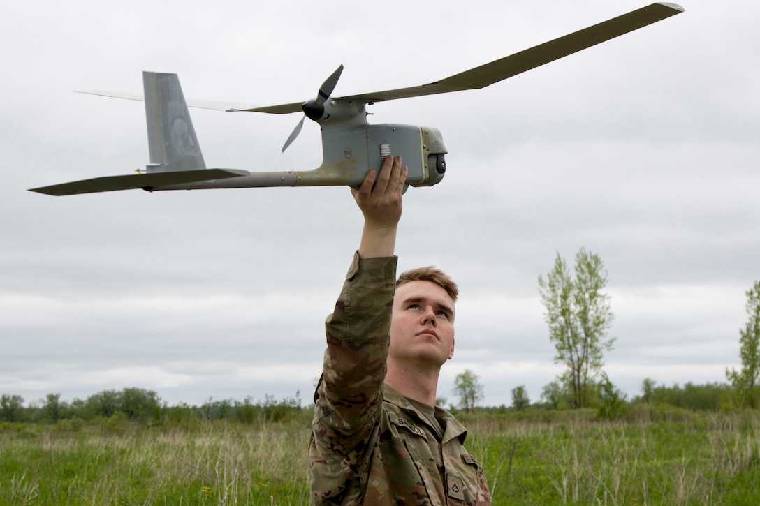 A soldier holds an unmanned drone aloft to inspect it in front of an open field.