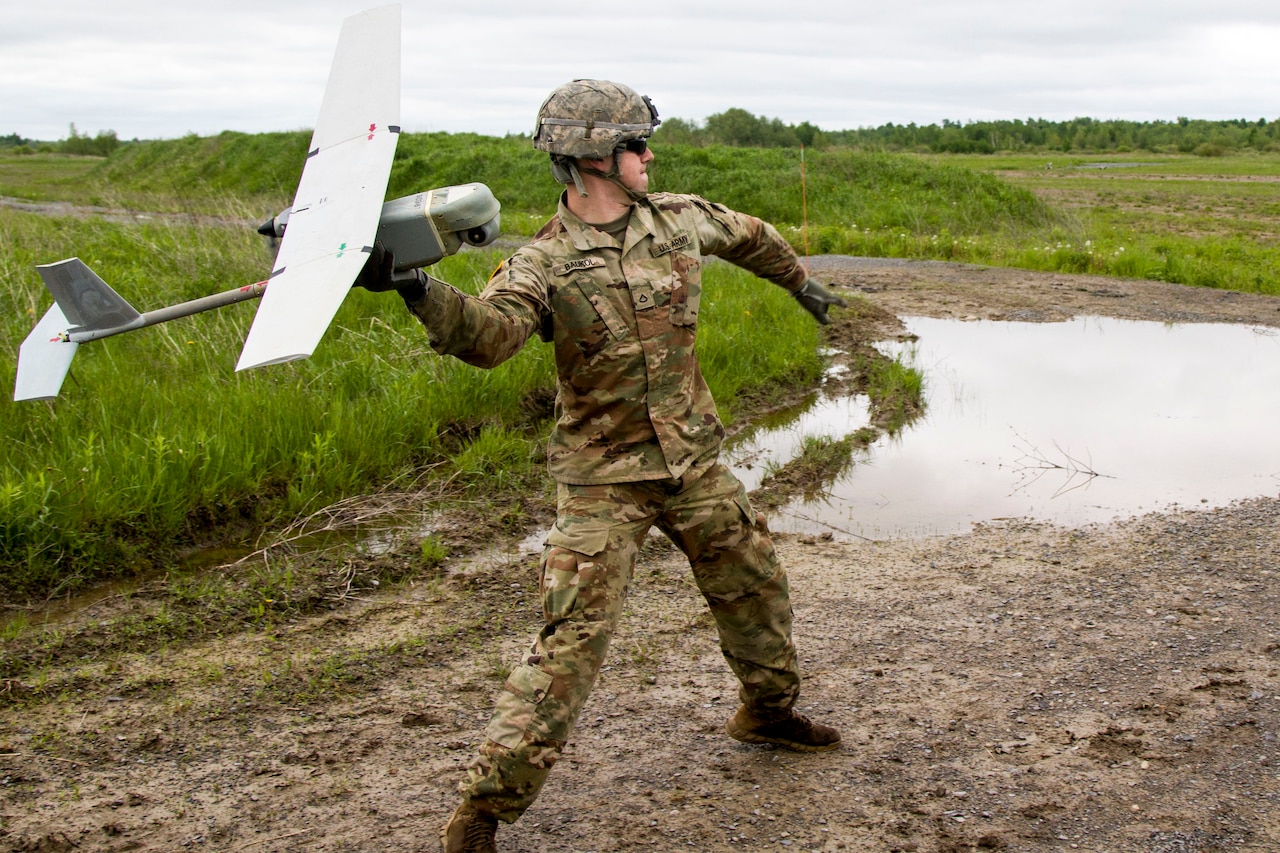 A soldier standing on a trail in a camouflage uniform, helmet and sunglasses winds up to release an unmanned drone into the air.