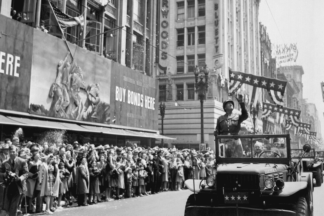 Army Gen. George S. Patton stands and waves from an open jeep as people line a downtown street.