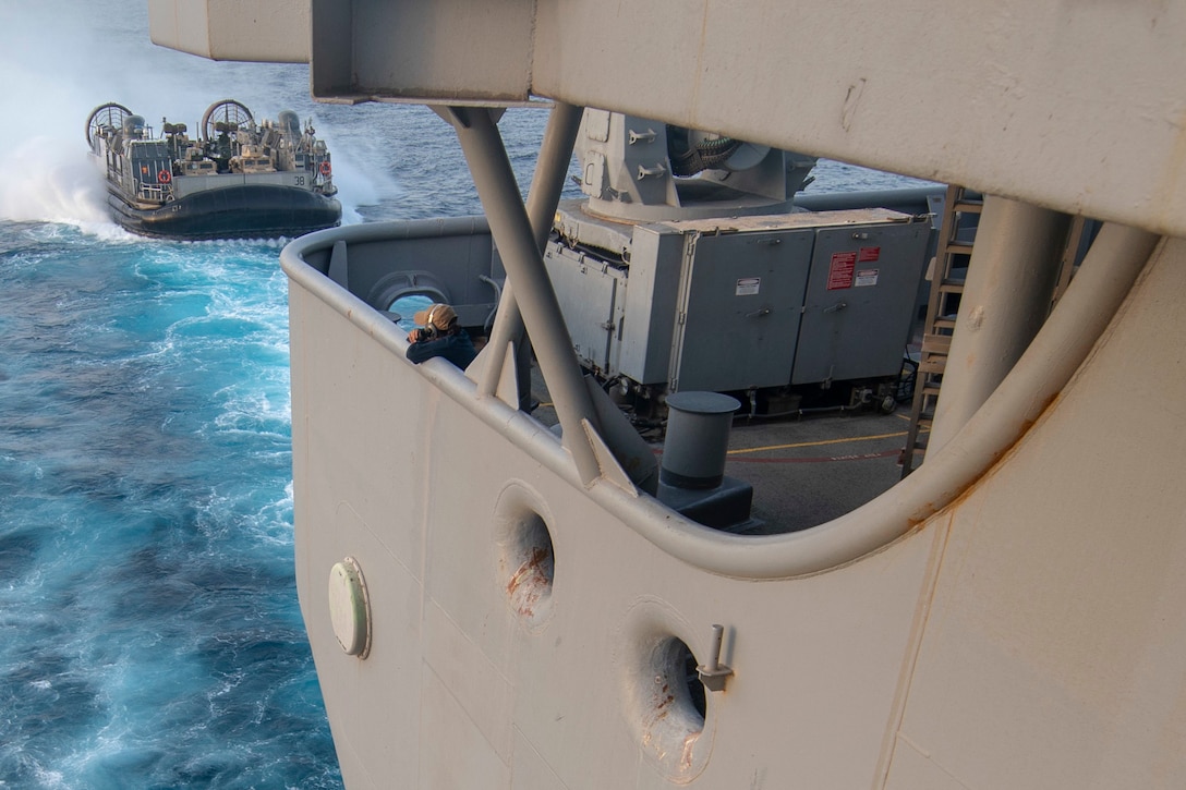 A hovercraft approaches the well deck of a ship as a sailor watches from above.
