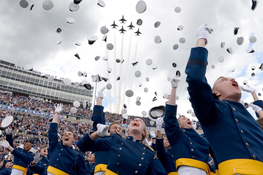 A group of graduates toss hats in the air as aircraft fly over a stadium.