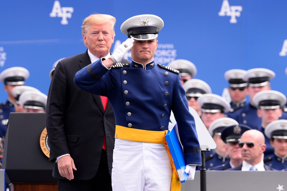 An Air Force cadet salutes on a stage as President Donald J. Trump looks on.