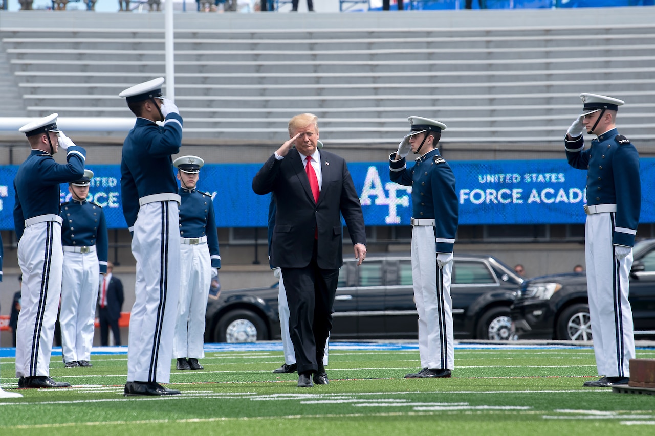 President Donald J. Trump salutes cadets as he walks on a football field.