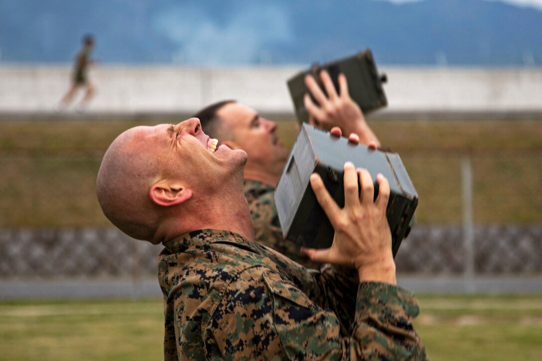 Two Marines lift ammunition cans above their chests while standing on an athletic field.