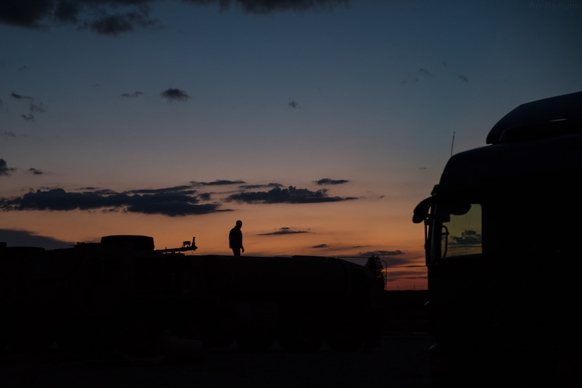 A service member stands in silhouette at dusk against an orange and blue sky.