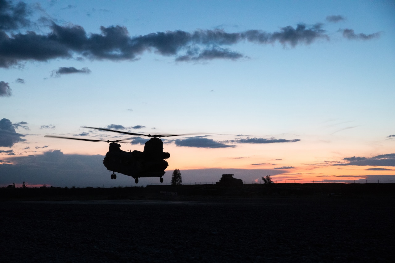 A helicopter with two rotors lifts off the ground in silhouette.