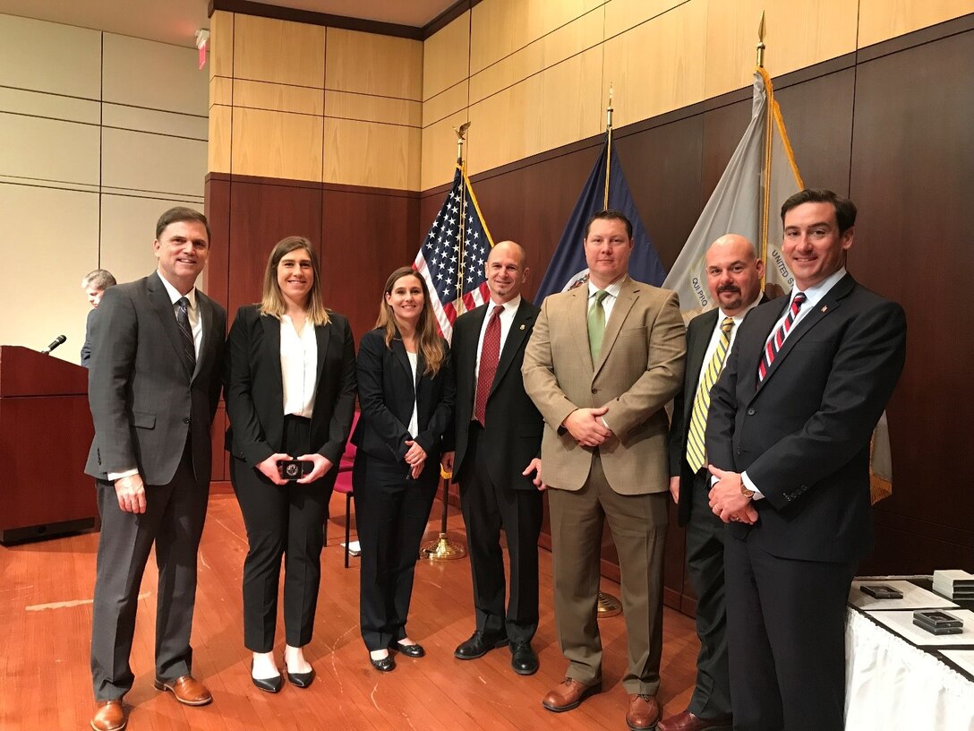 seven individuals stand in half circle with flags in background. award winner is second from left holder the award.