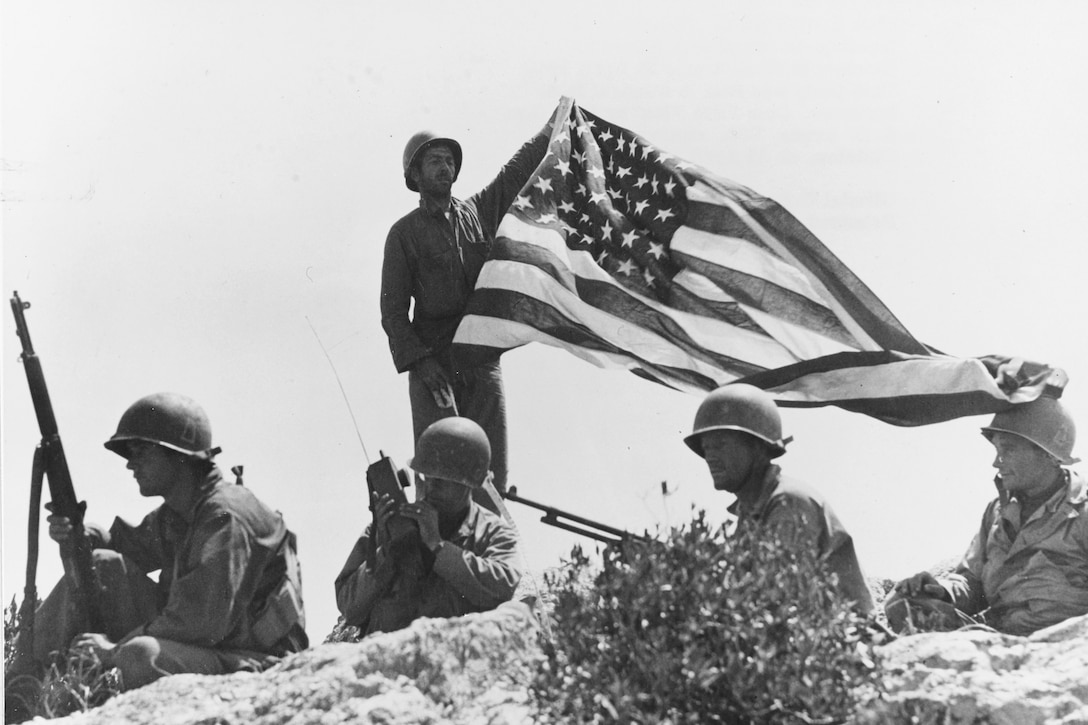 A soldier holds a flag up on a mountaintop as others crouch around him.