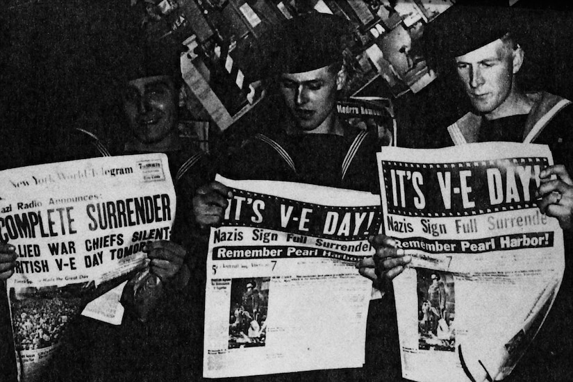 Three sailors read newspapers with giant 'It's V-E Day!' headlines.