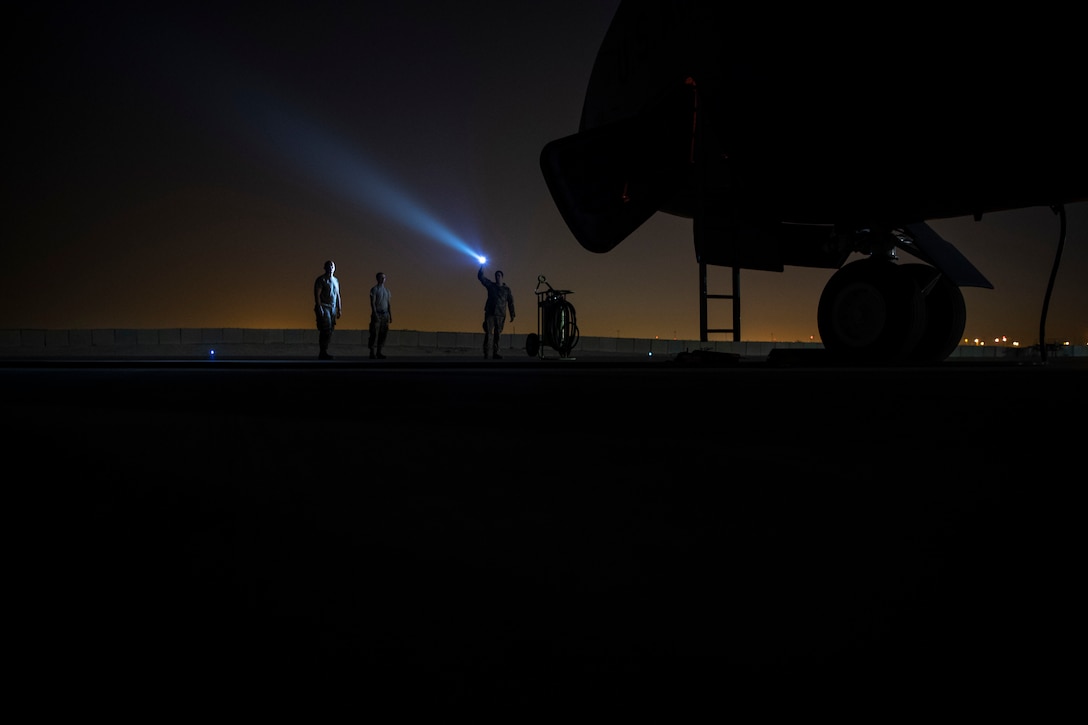 An airman shines a flashlight near a large aircraft at night as two other airmen look on.