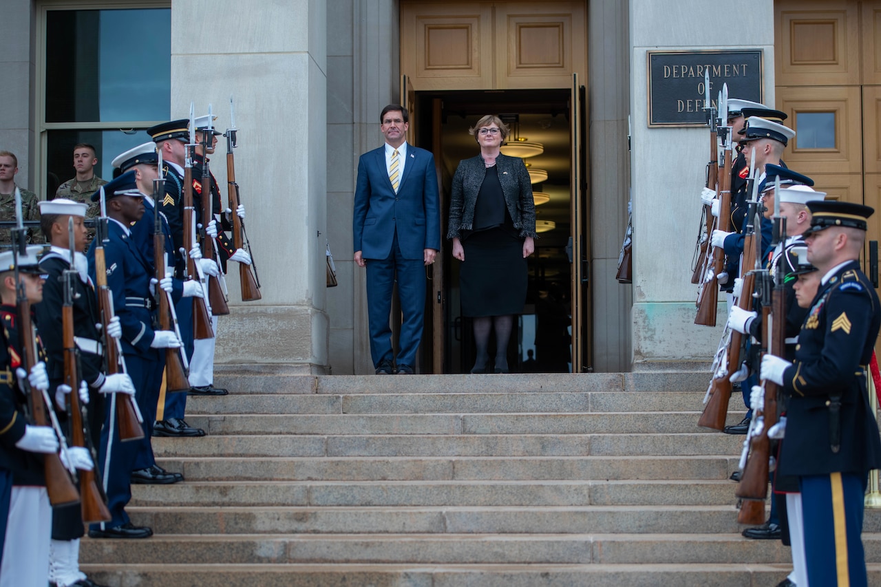 A man and woman stand at the top of steps, uniformed military personnel line the steps.