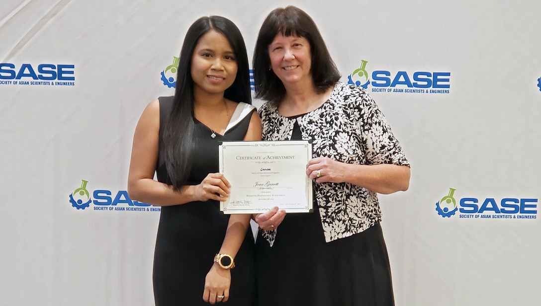 Two women stand in front of an event back drop holding an award.