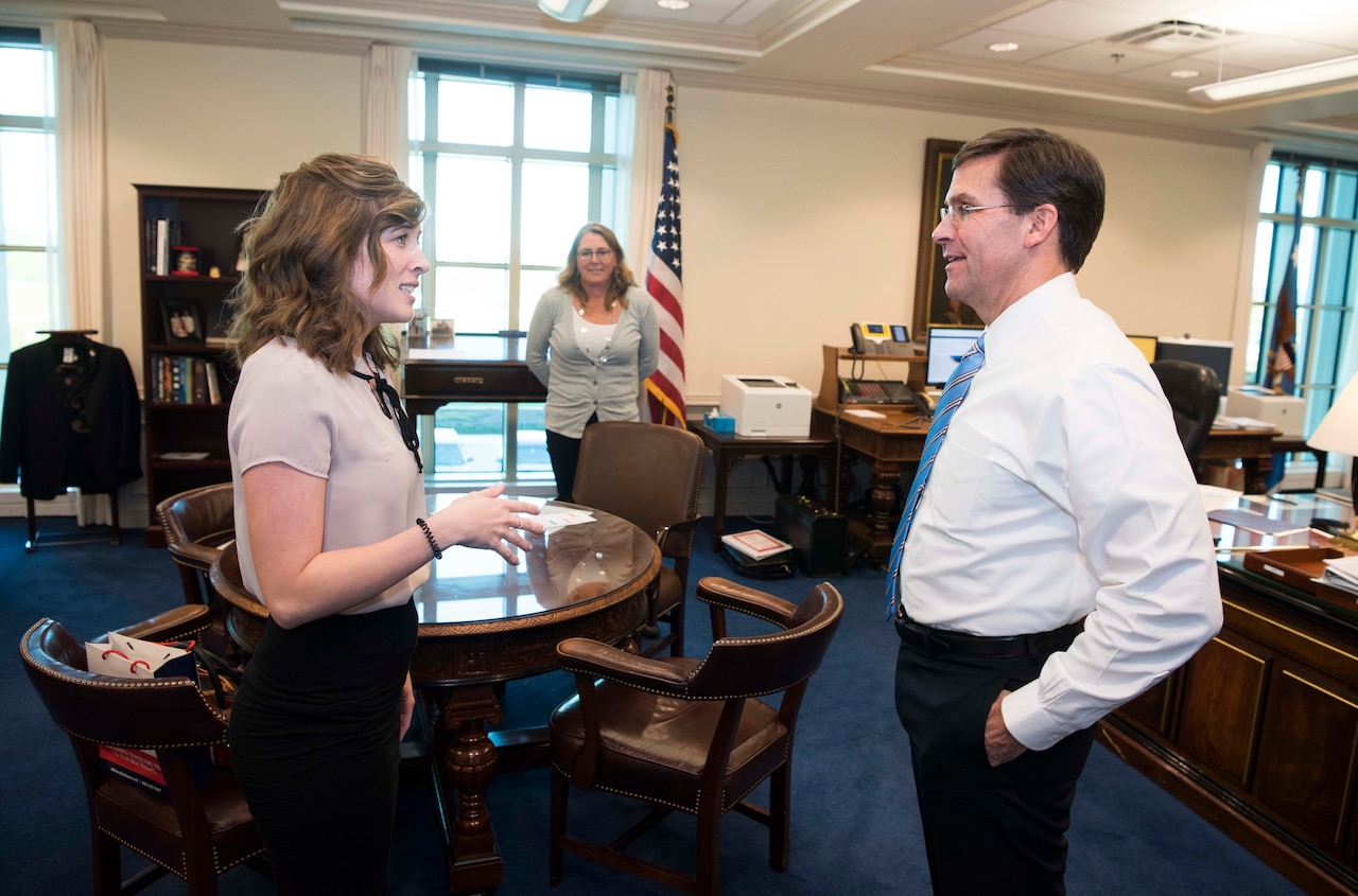 A woman and a man stand face to face and speak to each other in a large office. In the rear, another woman watches them.