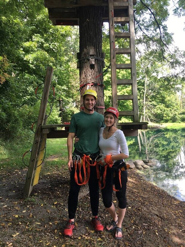 A man and woman wearing helmets stand outdoors in front of a tree. The tree has wooden platforms and ladders attached to its trunk.