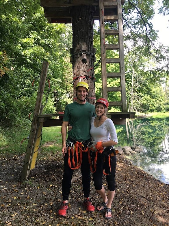 A man and woman wearing helmets stand outdoors in front of a tree. The tree has wooden platforms and ladders attached to its trunk.