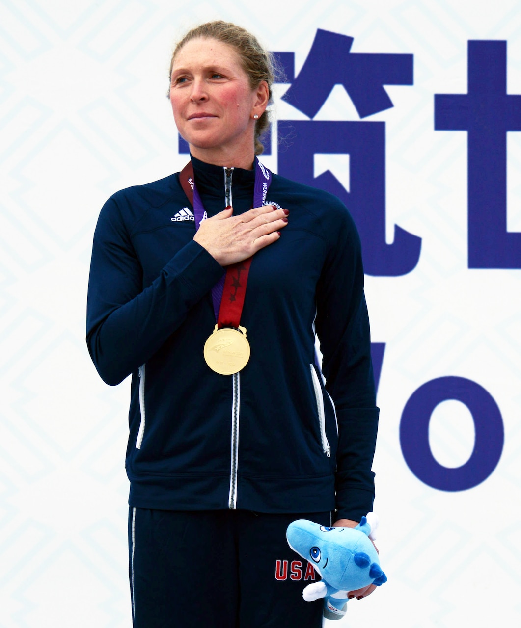 Woman wearing a blue track suit and a gold medal holds a stuffed animal in her left hand and holds her right hand over her heart.