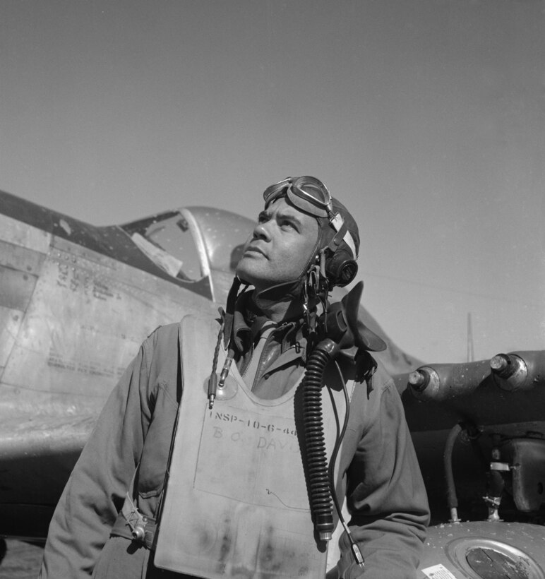 An airman looks at the sky while standing in front of a World War II-era aircraft.