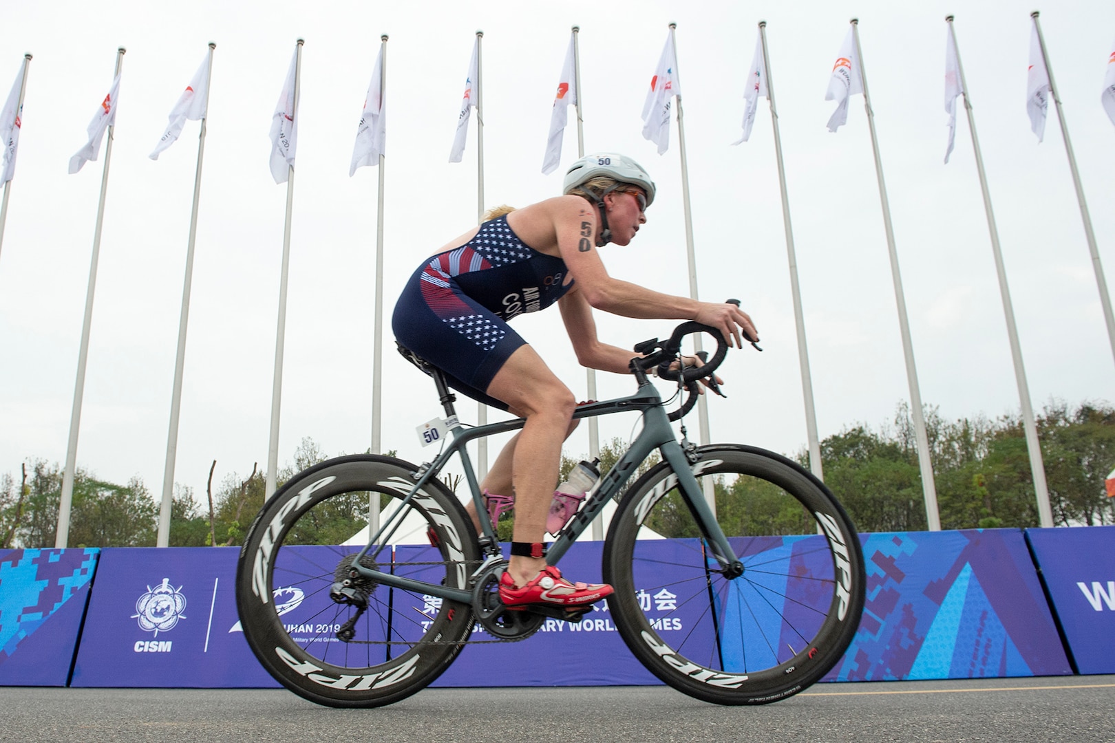 Woman triathlete rides a bicycle past a row of flagpoles that stand behind a blue course barrier.