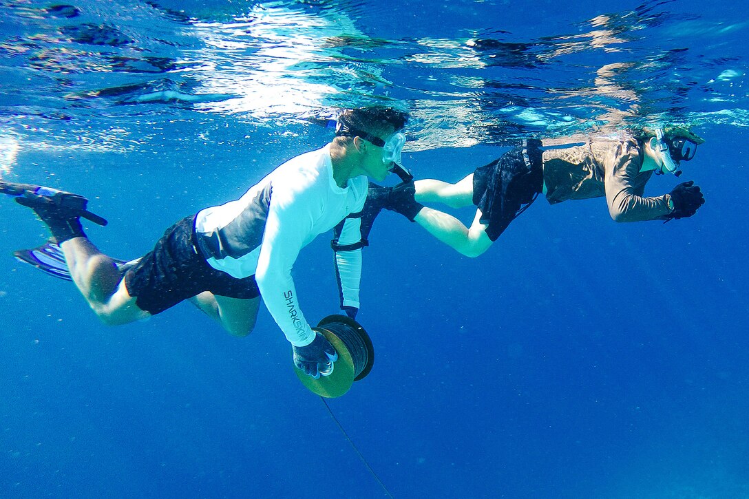 Two divers swim in vivid blue waters, one with a spool of detonation line.