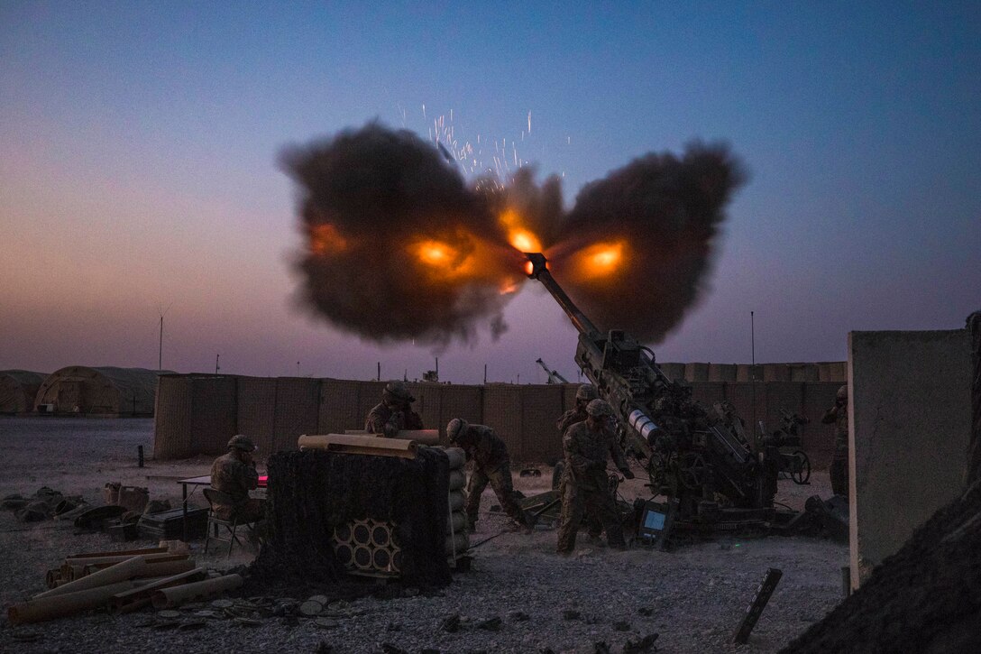 Soldiers fire a howitzer at an airfield, lighting up a dim blue sky.