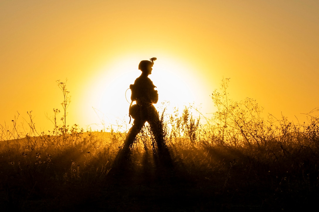 A Marine, shown in silhouette against a yellow-orange sky, walks across a field, the sun framing him from the background.