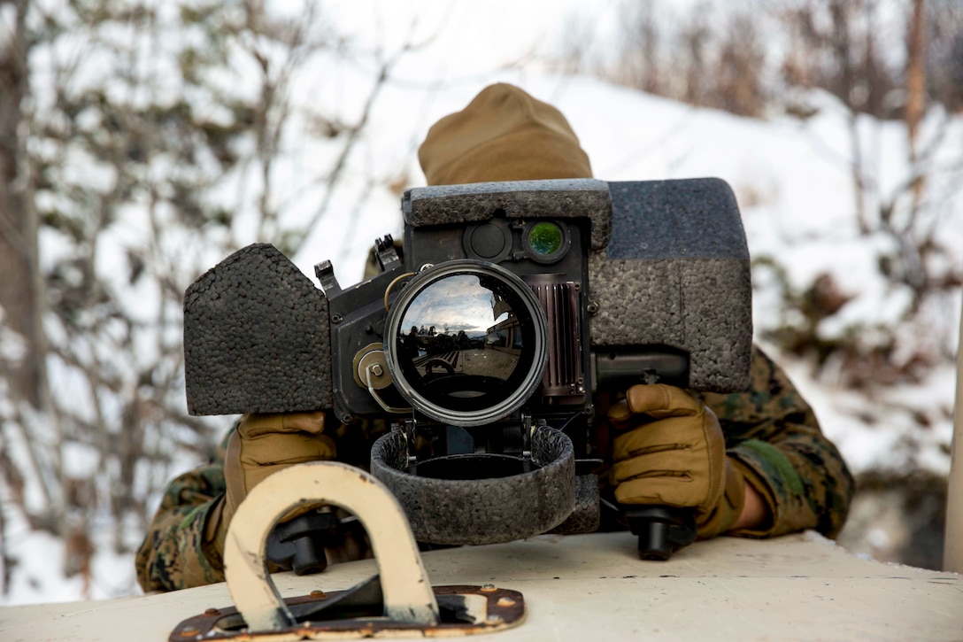 A Marine looks through a large lens in a snowy, wooded area.