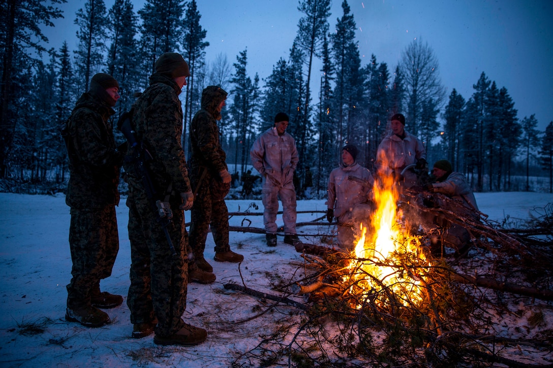 U.S. Marines and Norwegian soldier stand around a campfire on snowy ground, in low light.