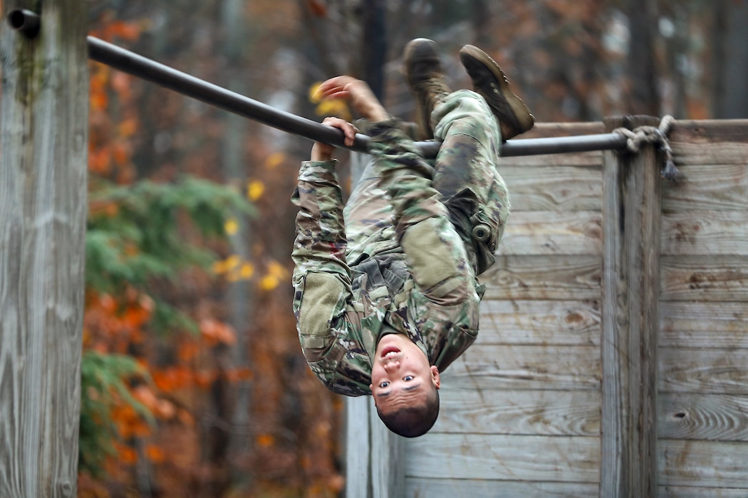 A soldier hangs by his arms and legs from a bar in a woodsy area.