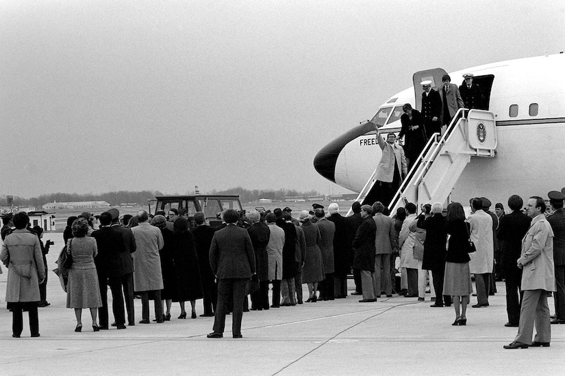 A group of people stand outside an airplane as people deboard.