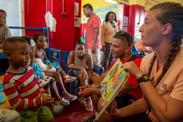 Two sailors read to a group of children in a red-painted room.
